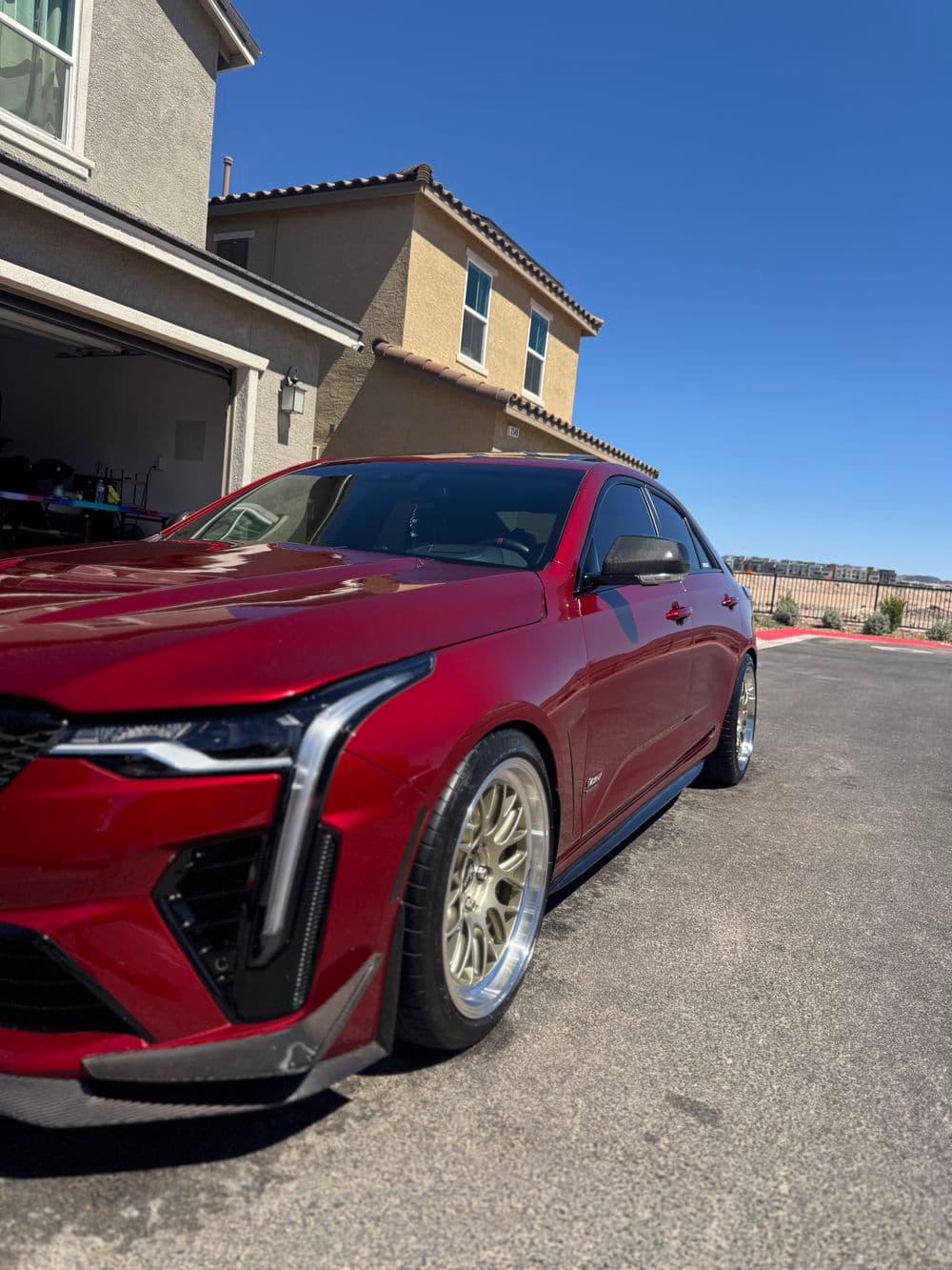 Red luxury sedan with gold wheels parked outside a house under a clear blue sky.